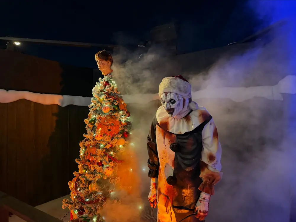 Art the Clown stands beside a burning, corpse-decorated tree in a smoke-filled scene from Terrifier at Halloween Horror Nights.