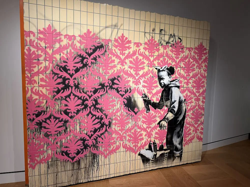 Banksy artwork featuring a young graffiti artist painting an ornate pink pattern, displayed at the Lucas Museum of Narrative Art.
