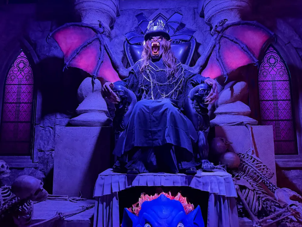A costumed figure with towering dark wings sits on an ornate stone throne under purple and blue light at Universal's Halloween Horror Nights.