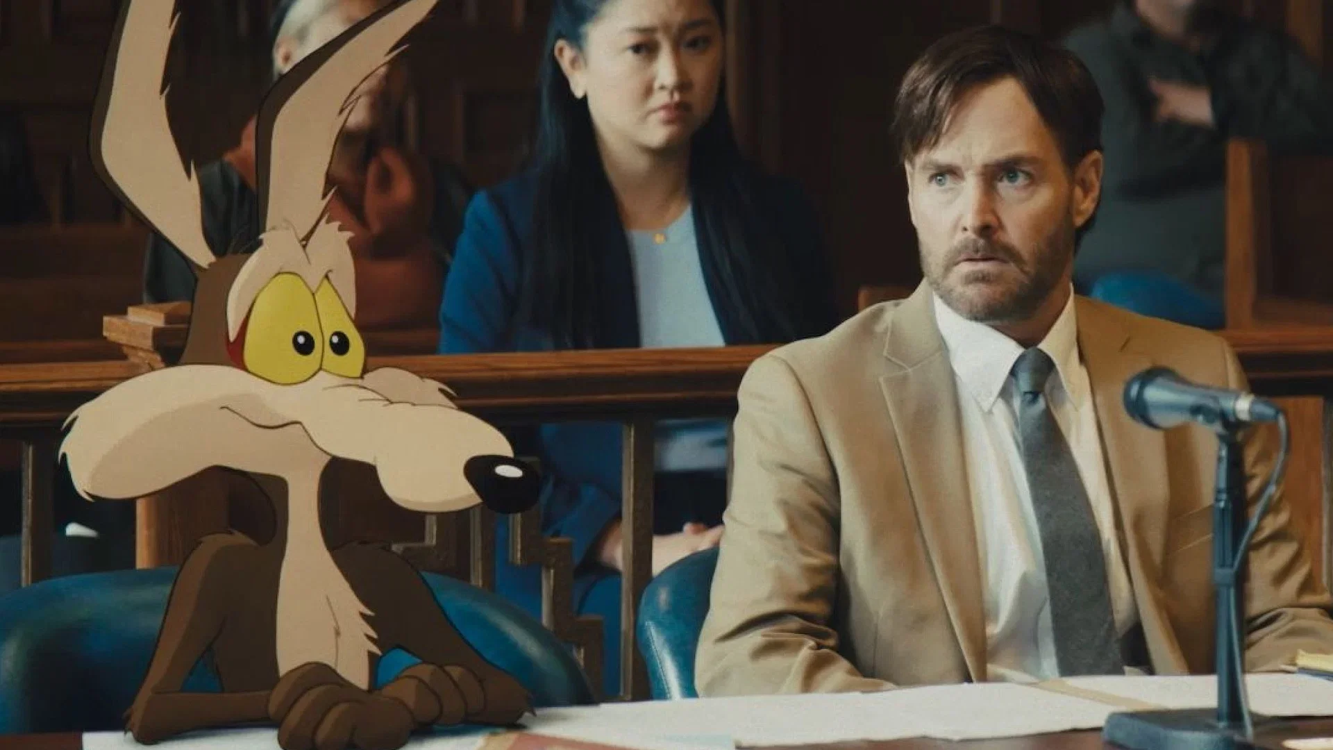 Wile E. Coyote sits at a courtroom table beside live-action actors in a scene from Coyote vs. Acme