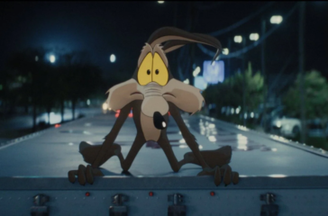 Wile E. Coyote sits at a courtroom table beside live-action actors in a scene from Coyote vs. Acme