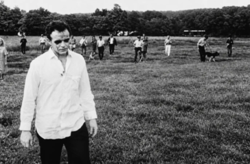 A ghoul in a white shirt stands in a field as the armed posse and their dogs close in behind him in Night of the Living Dead (1968), one of the most influential low-budget horror films.
