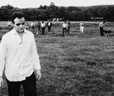 A ghoul in a white shirt stands in a field as the armed posse and their dogs close in behind him in Night of the Living Dead (1968), one of the most influential low-budget horror films.